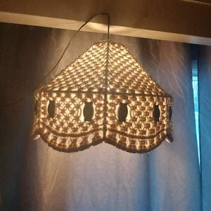 May include: A cream-colored macrame lampshade with a scalloped edge, hanging from a ceiling. The lampshade has a geometric pattern with oval cutouts. The light shines through the cutouts, illuminating the room. The lampshade is a decorative lighting fixture.