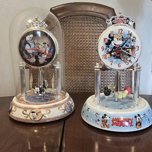 Può includere: Due orologi decorativi Disney. Un orologio presenta Pinocchio e Geppetto sotto una cupola di vetro. L'altro orologio presenta Topolino e i suoi amici. Entrambi gli orologi hanno dettagli argentati e sono su una base azzurra e bianca.