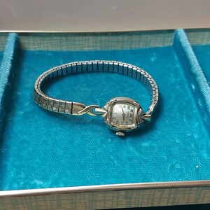 May include: A vintage Gruen quartz wristwatch with a silver-tone metal band and a rectangular watch face. The watch has a silver dial with black numerals and hands. The band is a stretch style, and the watch is set against a teal background.