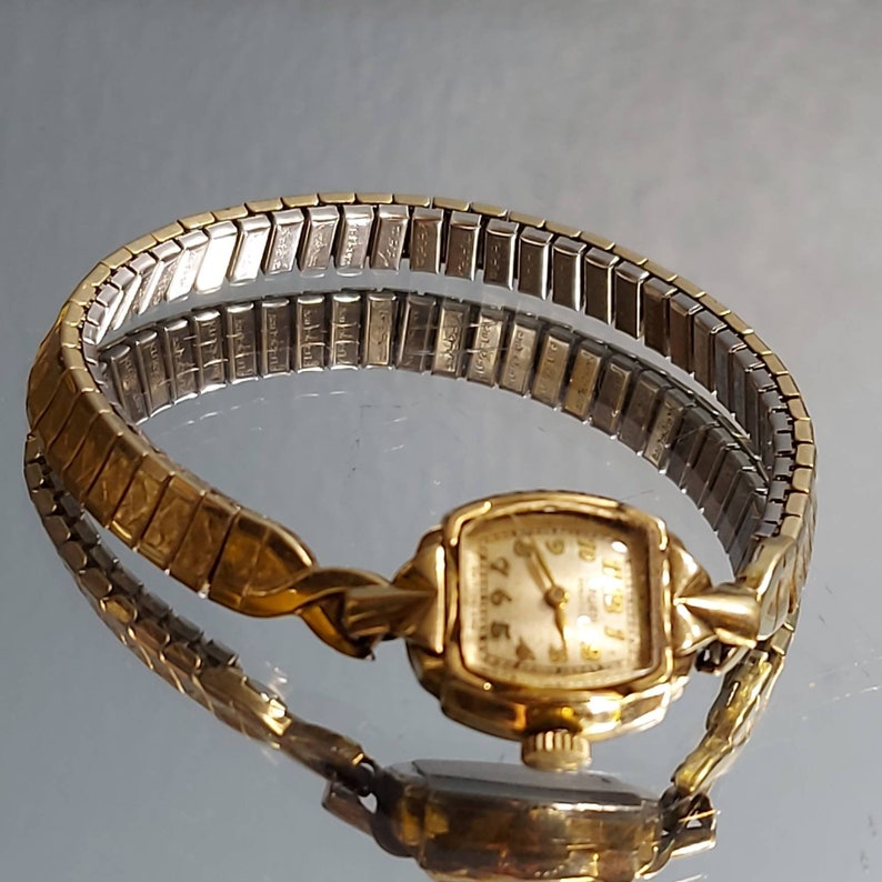 May include: A vintage gold-toned wristwatch with a rectangular face and a stretch band. The watch face has gold hands and numerals against a cream-colored background. The band is a mix of gold and silver-toned metal.