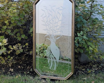 May include: An octagonal mirror with a gold-colored frame. The mirror's surface features an etched design of a vase with tall, slender plants. The mirror is displayed outdoors, resting on grass, with greenery in the background.