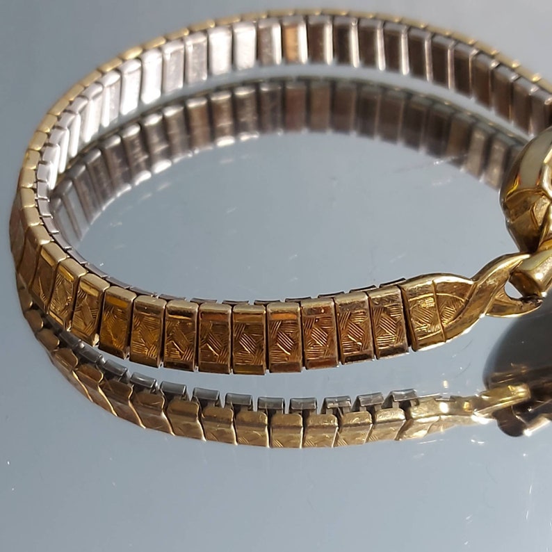 May include: A gold and silver-toned watch band with a rectangular link design. The gold links have a textured pattern. The band is reflected in a mirrored surface, showcasing its intricate design and craftsmanship. The watch band is curved.