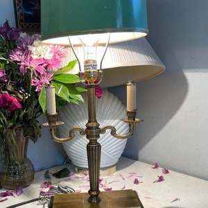 May include: A brass table lamp with a teal green lampshade and two candle-style light fixtures. The lamp has a rectangular base with electrical outlets. A vase of pink flowers and a white seashell-shaped object are in the background.