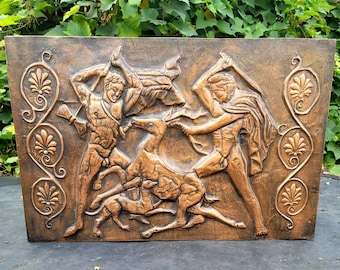 May include: A rectangular, bronze-colored relief sculpture depicting a hunting scene with two figures wielding weapons against a deer and dogs. The artwork features detailed figures and decorative floral elements along the sides.