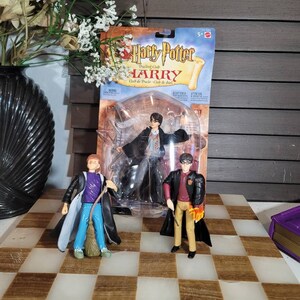 May include: A selection of Harry Potter action figures, including Harry, Ron, and another character, displayed on a checkered surface. The Harry figure is still in its original packaging. The figures are wearing robes and holding wands.