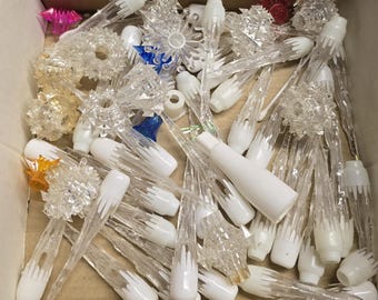 May include: A collection of assorted clear plastic icicle-shaped light covers and star-shaped ornaments. The pieces feature white plastic bases, some with faceted textures, and a few small accents in blue, orange, and pink, all loose in a box.