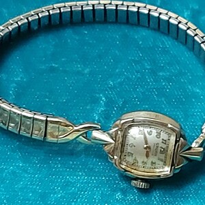 May include: Vintage Gruen watch with a silver-tone band and a gold-tone watch face. The watch face is square with a light-colored background and black numerals. The band is made of small, rectangular links.