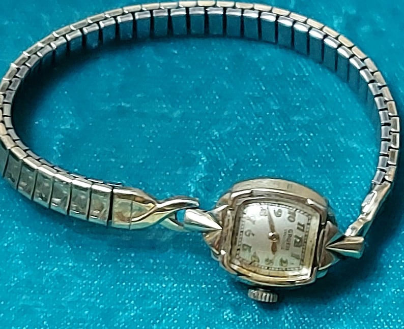 May include: Vintage Gruen watch with a silver-tone band and a gold-tone watch face. The watch face is square with a light-colored background and black numerals. The band is made of small, rectangular links.