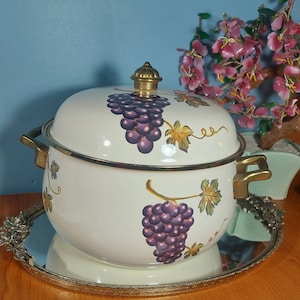Könnte beinhalten: Cremefarbener Emaille-Topf mit Deckel, verziert mit lila Trauben und goldenen Akzenten. Der Topf hat goldfarbene Griffe und einen goldenen Knauf. Er steht auf einem silbernen Tablett.