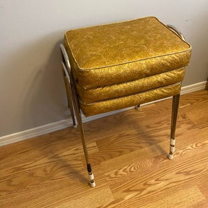 Puede incluir: Una caja de costura dorada vintage con un estampado floral. La caja tiene tres secciones apiladas con una parte superior acolchada y se apoya sobre delgadas patas de metal dorado con pies blancos. El marco es cromado.
