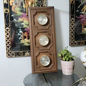 May include: A vintage wooden weather station with three circular gauges. The gauges have silver-coloured faces and black markings. The wooden frame is rectangular with decorative moulding. The weather station is displayed on a glass-topped table.