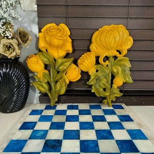 May include: Two yellow floral wall hangings, one a rose and the other a chrysanthemum, are displayed against a dark brown background. A blue and white checkered decorative piece is in the foreground. Decorative items.