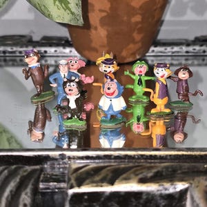 May include: A collection of eight small, colourful plastic figurines depicting various cartoon characters. The figures include animals and human-like characters in diverse outfits, such as a blue suit, a white jacket, and a green shirt, all standing on small green bases.