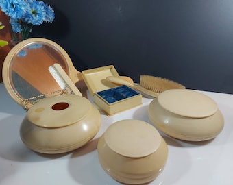 Può includere: Un set da toeletta vintage color crema, composto da uno specchio a mano, una spazzola per capelli, un pettine e diversi contenitori con coperchio. Il set include anche un piccolo portagioie con interno in velluto blu. Gli oggetti sono disposti su una superficie bianca.