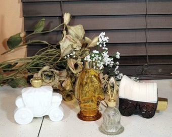 May include: A collection of vintage perfume bottles in various shapes. Includes a white carriage, a horse-shaped amber bottle, a clear bell, and a brown wagon. The bottles have gold-colored accents and are arranged on a white surface.