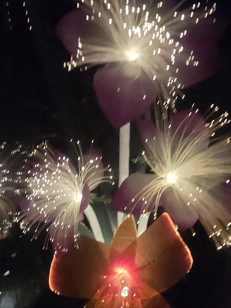 May include: Close-up of illuminated artificial flowers. The flowers have purple petals and white fiber optic filaments. The base of the flowers is orange, with a red glow from the center. The background is dark.