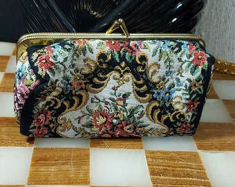 Puede incluir: Un monedero vintage de tapiz con cierre dorado. El bolso presenta un estampado floral en rojo, rosa, azul y verde sobre un fondo crema, enmarcado por un ribete negro. El cierre es de diseño rectangular.