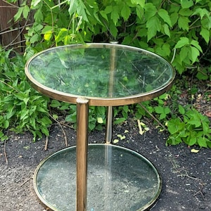 Puede incluir: Una mesa auxiliar de metal dorado con dos superficies circulares de vidrio. La mesa tiene un diseño elegante y moderno con una superficie superior y un estante inferior. Se apoya en tres patas doradas. El fondo presenta follaje verde.