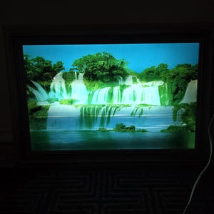 Puede incluir: Imagen enmarcada de una escena de cascada. La imagen muestra una cascada vibrante con follaje verde y cielo azul. El marco es oscuro, contrastando con la imagen brillante. La escena evoca una sensación de tranquilidad.