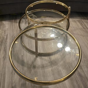Peut inclure: Un ensemble de deux tables basses gigognes avec des plateaux circulaires en verre transparent encadrés de métal doré poli. La structure se compose de deux tables rondes de hauteurs différentes, soutenues par des pieds verticaux fins en métal.