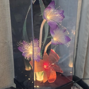 May include: A decorative lamp featuring artificial flowers encased in a clear, rectangular box. The flowers include purple blooms with fiber optic accents and an orange flower. The base is black.