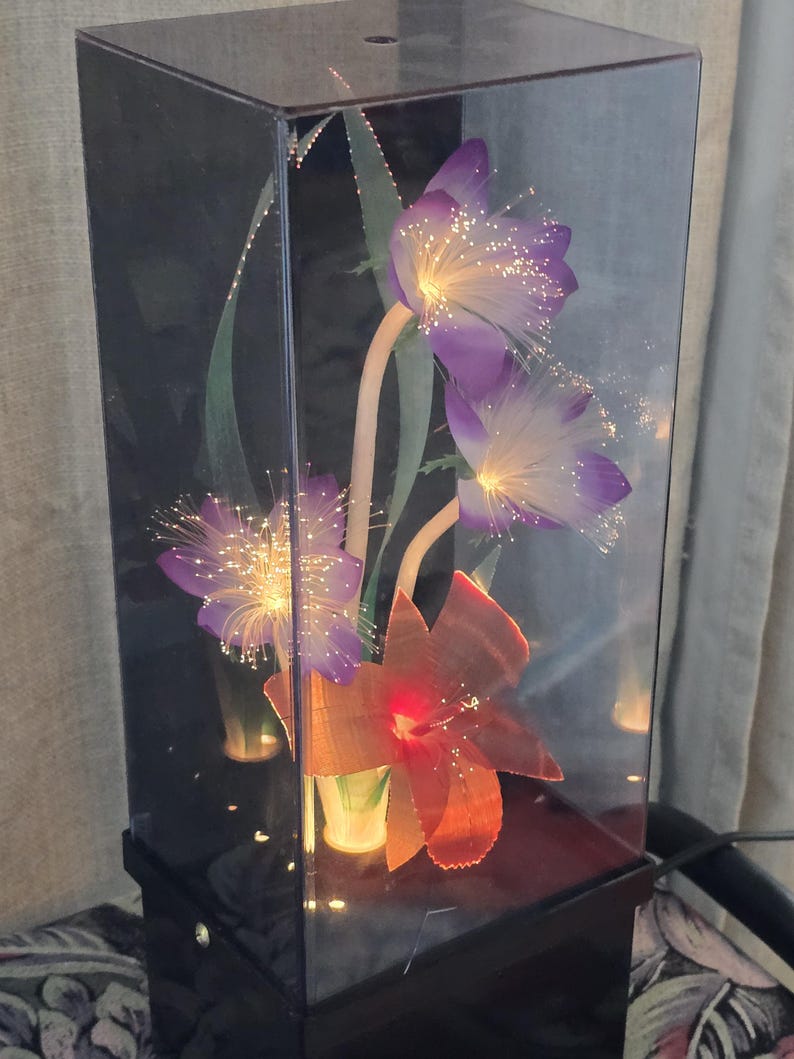 May include: A decorative lamp featuring artificial flowers encased in a clear, rectangular box. The flowers include purple blooms with fiber optic accents and an orange flower. The base is black.