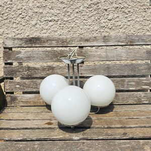 May include: A retro three-light chandelier featuring a chrome frame and three spherical, white glass shades. The light fitting is suspended from a circular ceiling plate. The chandelier is designed for interior lighting. The fixture is sitting on a wooden surface.