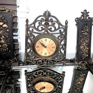 May include: Ornate, antique-style clock set with a central round clock and two side panels. The clock face is gold with Roman numerals. The frames are black with gold accents and decorative carvings. The set is likely made of metal.