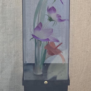May include: A decorative display case with artificial flowers. The flowers are purple and white, with green stems and leaves. The case has a black base and a clear, rectangular glass enclosure. The design is elegant and suitable for home decor.