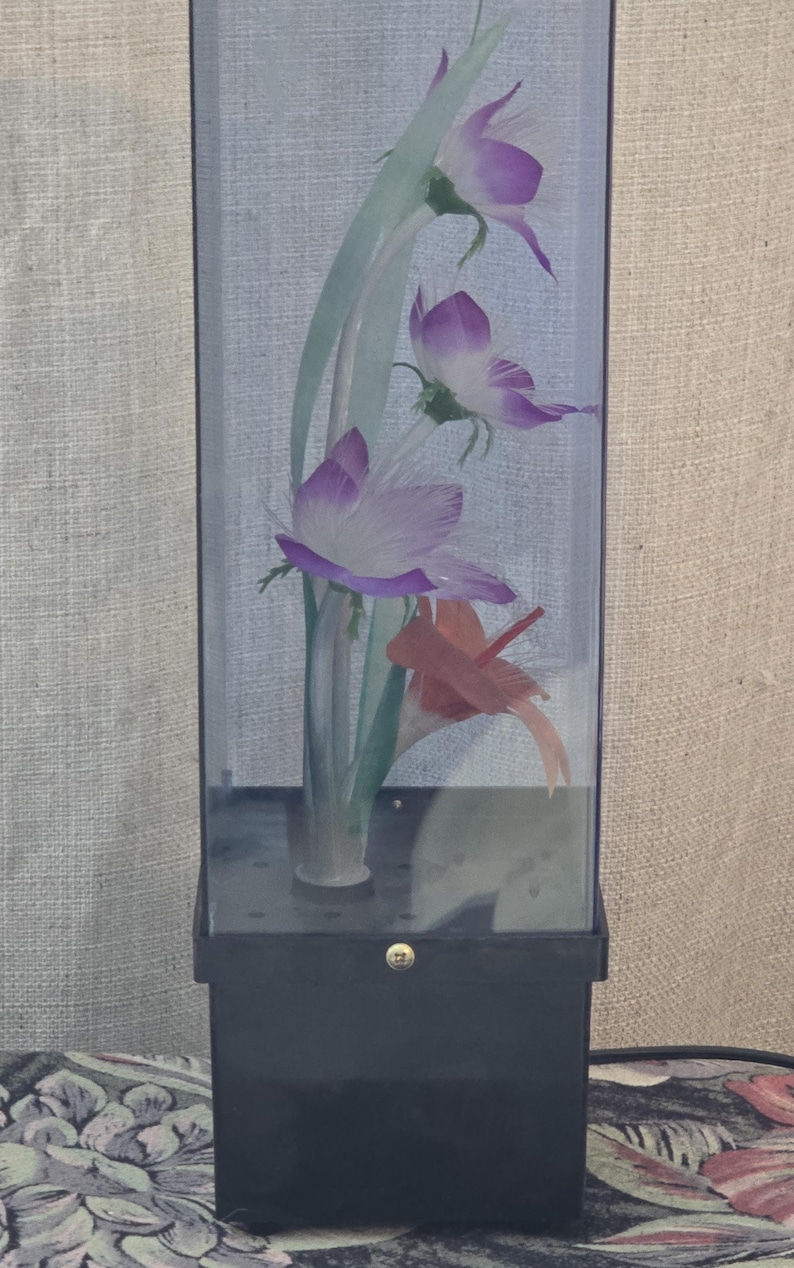 May include: A decorative display case with artificial flowers. The flowers are purple and white, with green stems and leaves. The case has a black base and a clear, rectangular glass enclosure. The design is elegant and suitable for home decor.