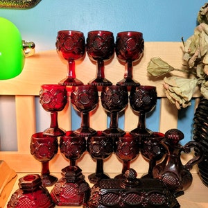 Op de afbeelding: Een verzameling robijnrood glaswerk, waaronder wijnglazen, een kan en een botervloot. De glazen hebben een gedetailleerd patroon. De items zijn gerangschikt op een houten oppervlak, wat een vintage esthetiek creëert.