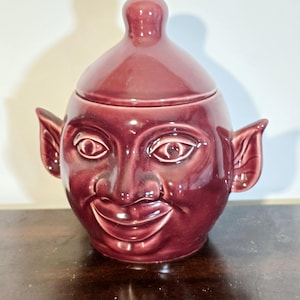 Könnte beinhalten: Eine bordeauxfarbene Keramikdose mit Deckel, die ein Gesicht mit großen Augen, einem lächelnden Mund und spitzen Ohren zeigt. Die Dose hat eine bauchige Form und einen kleinen Griff auf dem Deckel. Das Design ist skurril und dekorativ.