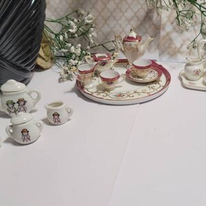 Puede incluir: Juegos de té de porcelana en miniatura. Un juego presenta una tetera, una crema, un azucarero y tazas con un diseño de niños. Otro juego tiene un diseño floral con una bandeja. Los juegos son blancos con detalles en rojo y dorado.
