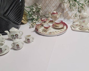 May include: Miniature porcelain tea sets. One set features a teapot, creamer, sugar bowl, and cups with a design of children. Another set has a floral design with a serving tray. The sets are white with red and gold accents.