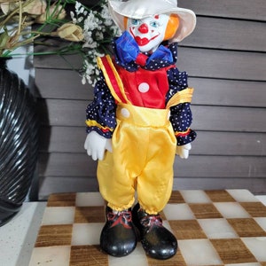 May include: A porcelain clown doll with a white hat, orange hair, and a painted red smile. The doll wears a red and blue outfit with yellow trousers and black shoes. The doll is standing on a checkered surface.