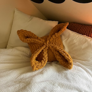 May include: A mustard yellow knitted throw pillow in the shape of a four-pointed star. The pillow is on a white bed with a white pillow behind it.