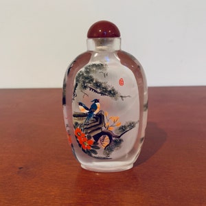 May include: A clear glass bottle with a red stopper and a painted scene of two birds perched on a branch with flowers. The bottle is oval shaped and has a frosted finish.