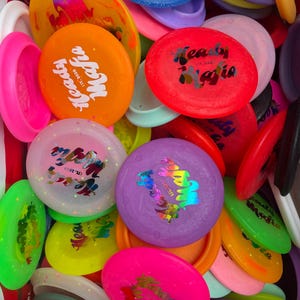 Pode incluir: Uma pilha de frisbees coloridos com as palavras "Heady Mafia" e "Headie Mafia" impressas neles. Os frisbees são de várias cores, incluindo vermelho, laranja, amarelo, verde, azul, roxo e rosa.