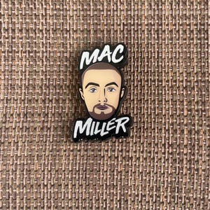 May include: A black and white shoe charm featuring a cartoon illustration of Mac Miller's face. The charm has the name "MAC" above the face and "MILLER" below it, both in white lettering.