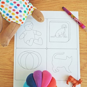 May include: A white sheet of paper with four dotted line drawings of a person, a dog, a pumpkin, and a cat. A rainbow-colored fabric ball and a small orange toy cat are on the paper.