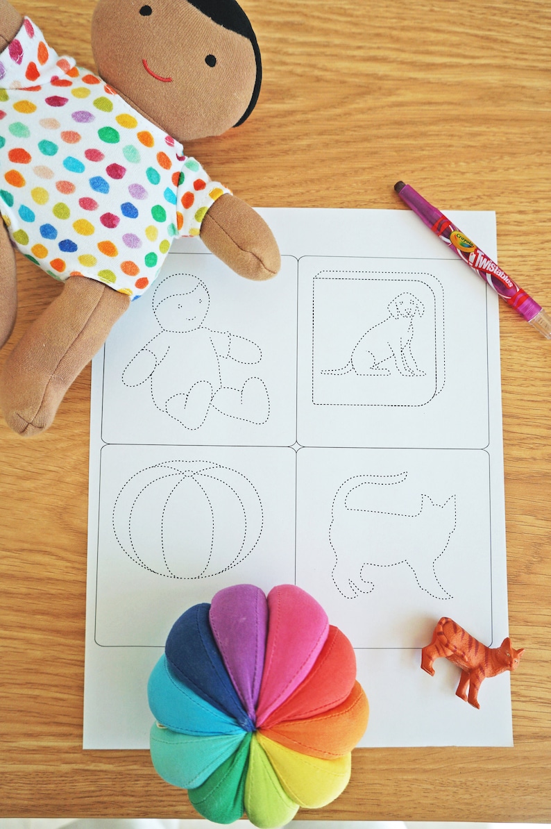 May include: A white sheet of paper with four dotted line drawings of a person, a dog, a pumpkin, and a cat. A rainbow-colored fabric ball and a small orange toy cat are on the paper.