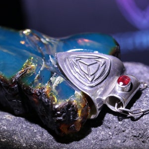 Puede incluir: Colgante de ámbar azul con engaste de plata. El colgante presenta un diseño geométrico y una pequeña gema roja. El ámbar tiene una textura natural y rugosa, sobre un fondo oscuro.