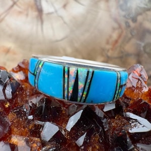 May include: A silver ring with a turquoise band, accented with black and iridescent inlays. The ring is set against a backdrop of dark, sparkling crystals, creating a striking contrast of colors and textures. The ring's design is geometric and modern.