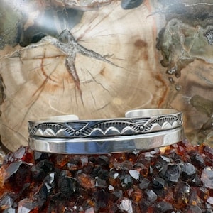 May include: A silver cuff bracelet featuring a polished lower band and an upper band decorated with engraved geometric patterns, including repeating triangles and small circular accents, with an open back design for adjustable fit.