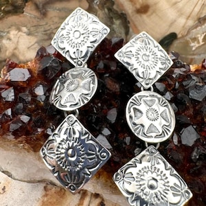 May include: A pair of silver-toned drop earrings featuring a vertical arrangement of alternating diamond and circular shapes. Each metal component is embossed with intricate, symmetrical floral-inspired patterns and finished with a polished surface.
