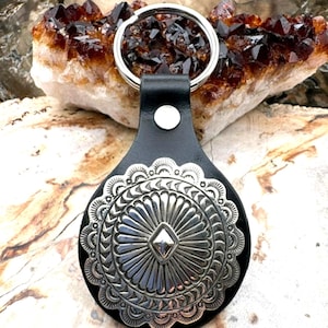 May include: Black leather keychain with a large, silver-colored decorative medallion. The medallion features an intricate, detailed design with a central diamond shape and a scalloped edge. A silver keyring is attached to the leather.