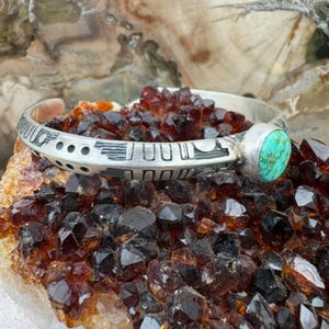May include: A silver cuff bracelet featuring a round turquoise stone set at one end. The metal band displays a series of etched geometric patterns, including vertical lines and circular indentations, creating a textured surface along the exterior.