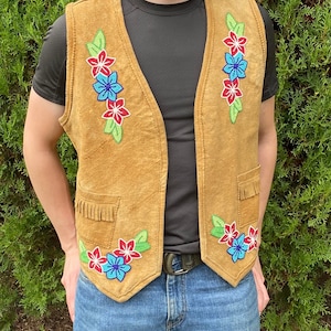 May include: Tan suede vest with floral beadwork in red, blue, and white. The vest has a V-neck, two front pockets with fringe, and a decorative floral pattern on the front. The vest is a medium size.