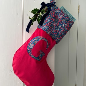 May include: A bright pink Christmas stocking featuring a floral patterned cuff and a matching letter G appliqué. The stocking is adorned with a dark blue velvet ribbon bow and a sprig of holly, with a light blue book tucked inside the top opening.
