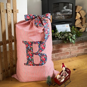 May include: A large red and white striped gift sack with a floral letter "B" appliqué and a matching bow. A wooden sled and a small toy mouse in a sleigh are in the foreground. A decorative bow sits atop a black stove.